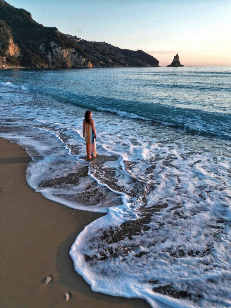 Spacerująca kobieta na plaży Glyfada na Korfu o zachodzie słońca, z widokiem na fale i skały